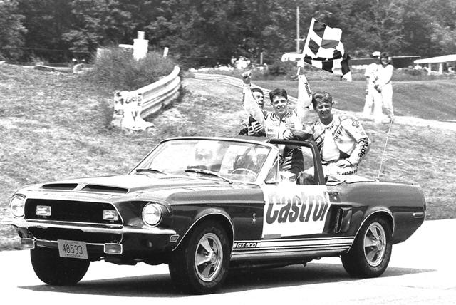 A victory lap for the podium finishers after the 1989 Road America AMA Castrol 250 Grand Prix race. The winner holding up the flag is John Kocinski. He is flanked by second-place finisher Rich Oliver (behind Kocinski) and third-place Alan Carter (with the Aprilia leathers). (Larry Lawrence photo)